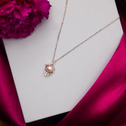 A silver pendant chain featuring a leaf design with a pearl in the center, displayed on a white surface with a pink flower and satin fabric in the background.